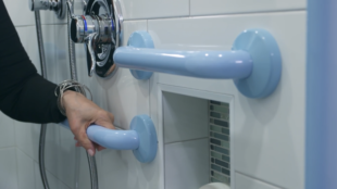 The image shows a close-up of a person's hand gripping a blue grab bar in a bathroom setting. The grab bar is mounted on a white tiled wall, next to a silver shower unit that has a chrome finish. There is a shower hose attached to the unit, and below the grab bar, there is a recessed niche in the wall that contains a soap dish with a bar of soap.